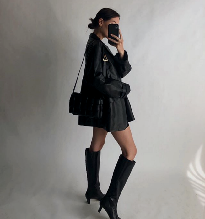 chica de cabello oscuro usando un saco de cuero negro, minifalda negra, top negro, botas largas de tacón y bolso negro pequeño