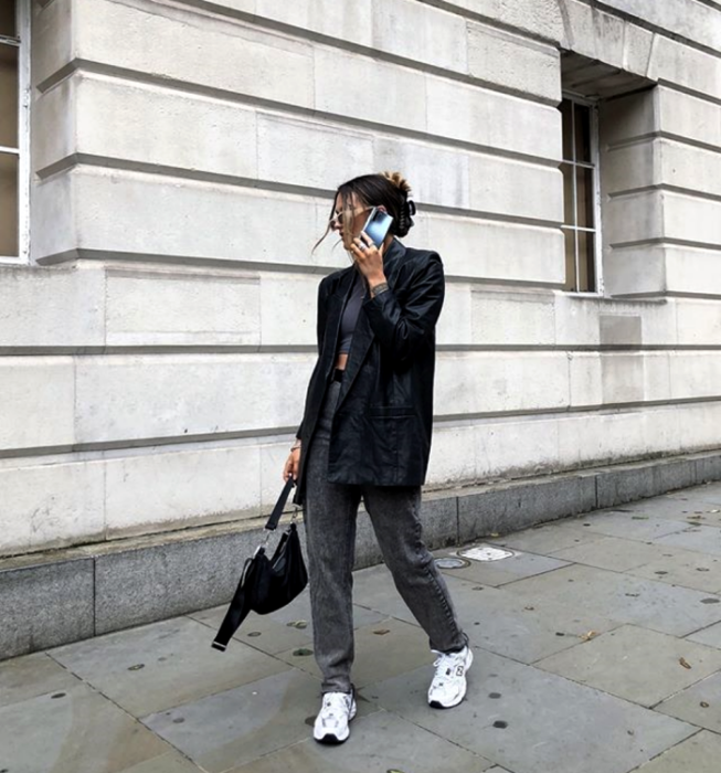 braunhaariges Mädchen, das ein graues Crop-Top, einen langen Ledermantel, graue Jeans, weiße Tennisschuhe und eine schwarze Handtasche trägt
