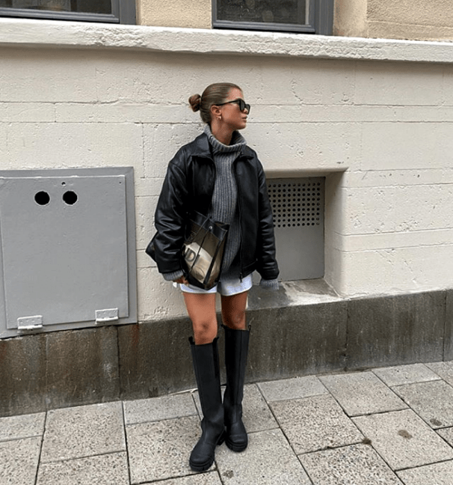 chica de cabello castaño usando lentes de sol, suéter tejido gris de cuello alto, saco de cuero, shorts blancos, botas largas de cuero de suela gruesa y bolso de mano semitransparente