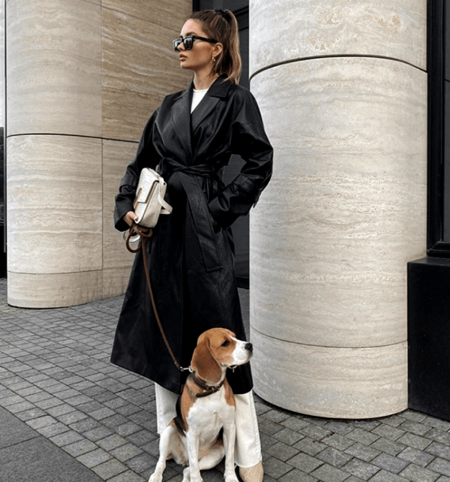 chica de cabello claro usando lentes de sol, abrigo largo de cuero, camiseta blanca, pantalones blancos de vestir, botas beige de cuero y bolso de mano blanco