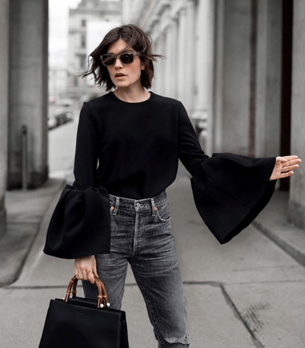 chica de cabello corto usando lentes de sol, blusa negra con mangas abombadas, jeans a la cintura y bolso de mano negro con correa de bambú