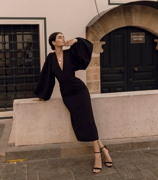 chica de cabello oscuro usando un vestido negro ceñido, con mangas abombadas, escote en V y sandalias de tacón negras