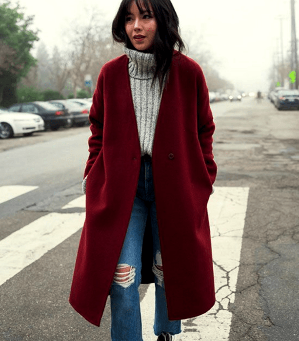 chica de cabello castaño usando un suéter gris tejido de cuello alto, abrigo largo rojo, jeans ajustados rotos de la rodilla, botines negros de tacón