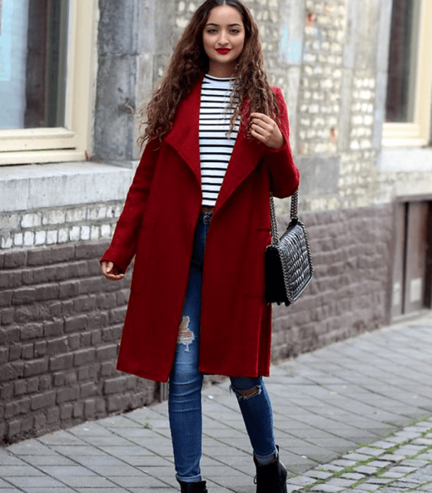 chica de cabello castaño largo usando un top blanco de rayas negras, abrigo largo rojo, skinny jeans, botines negros de piel y bolso de mano  negro