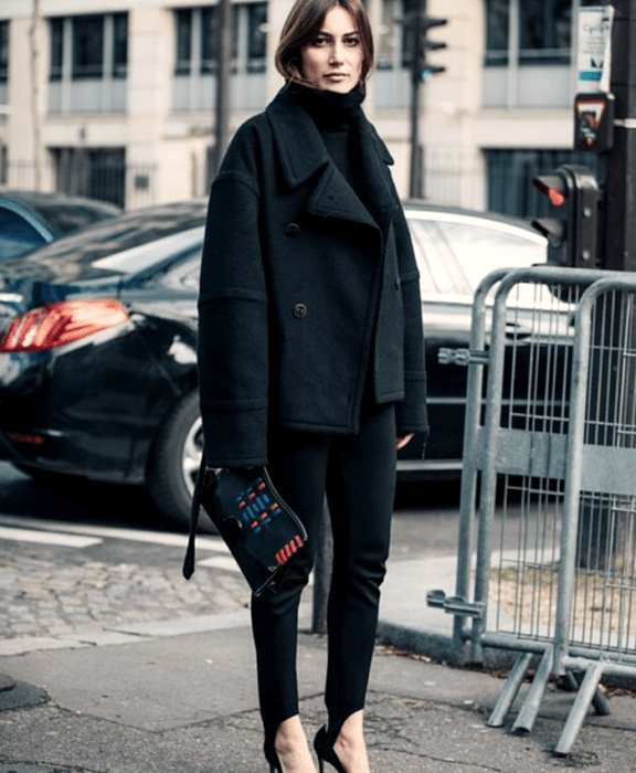 chica de cabello castaño usando un suéter de cuelo alto negro, abrigo grande negro, leggings con estribos negros, zapatos de tacón y bolso de mano negro con vistas rojas y azules