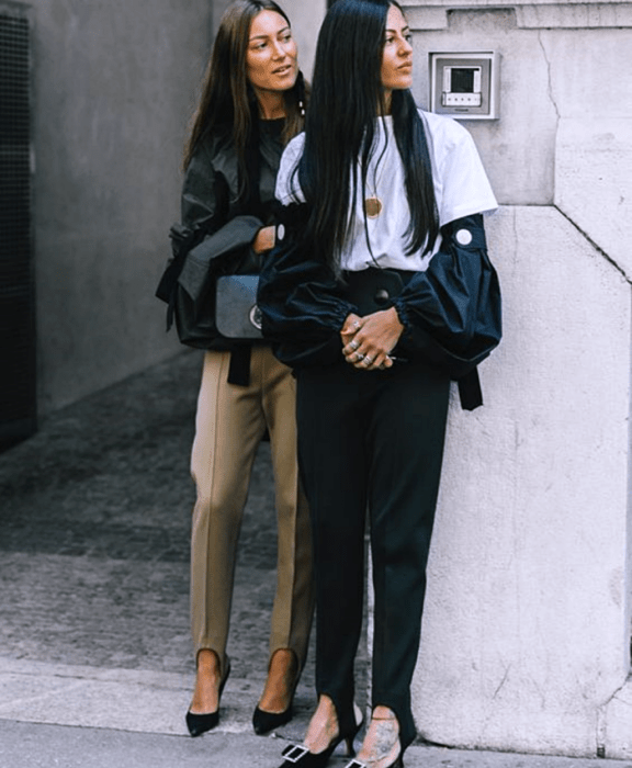 dos chicas de cabello largo usando una camiseta blanca, top negro, leggings negros y cafés con estribos, zapatos de tacón negro, chaqueta negra y bolso negro de mano