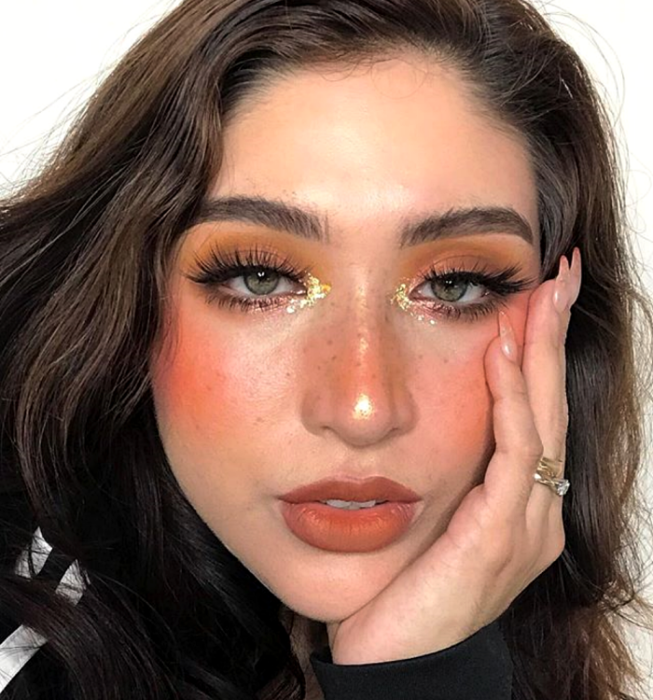 chica de cabello castaño usando una sombra naranja, pestañas postizas, blush intenso, lipstick naranja oscuro y brillo glitter dorado en el lagrimal