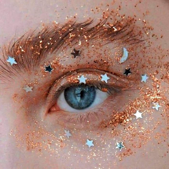 maquillaje con sombras brillantes naranjas, glitter y stickers plateados en forma de estrella y lunas
