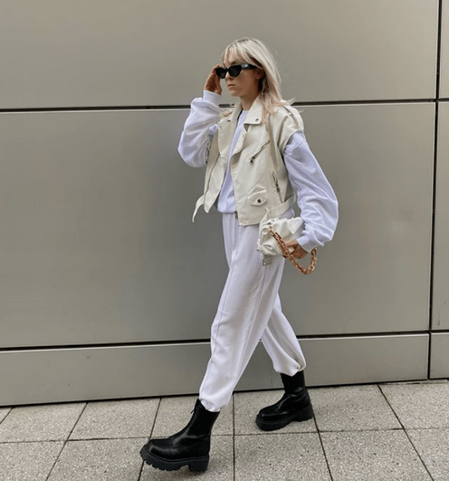 chica rubia platinada usando lentes de sol, sudadera blanca, chaleco de mezclilla blanco, pants blancos, botas negras de suela gruesa y bolso de mano beige