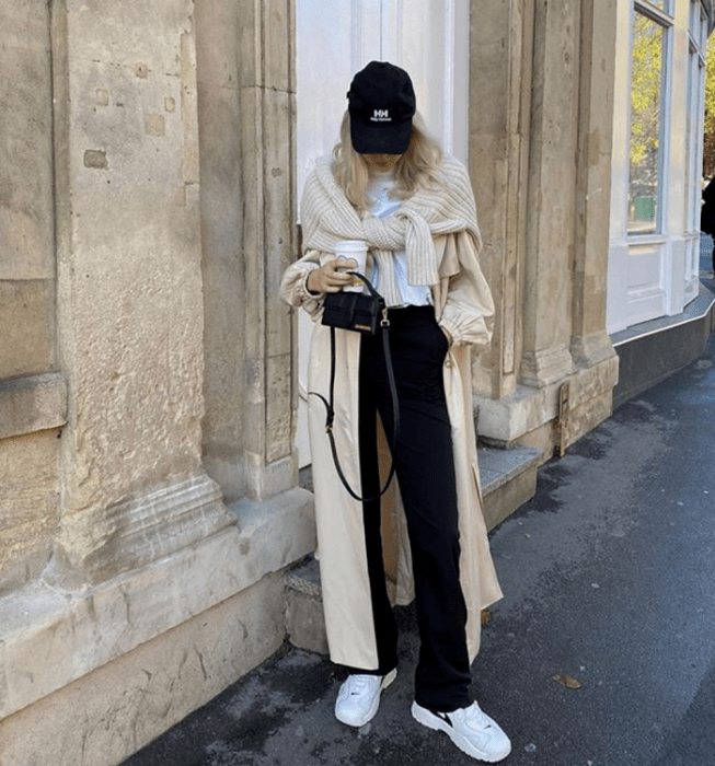 chica rubia usando gorra negra, camiseta blanca, pants negros, tenis blancos, abrigo largo beige, suéter beige tejido y bolso de mano mini negro