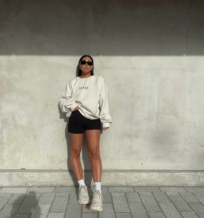 chica de cabello castaño usando lentes de sol, sudadera blanca, shorts de ciclismo negros, tenis blancos