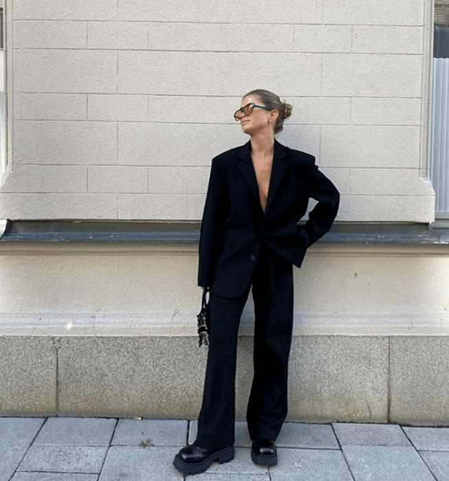 chica rubia usando lentes de sol, blazer negro, pantalones negros de vestir, botas negras de cuero con plataforma y suela gruesa y bolso negro mini