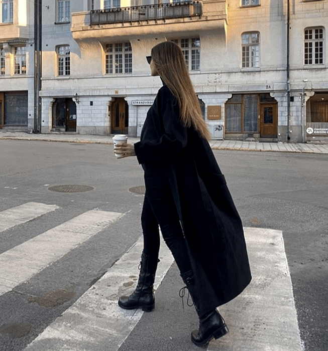 chica rubia usando lentes de sol, abrigo largo negro, skinny jeans negros, botines negros de plataforma y top  negro de cuello alto