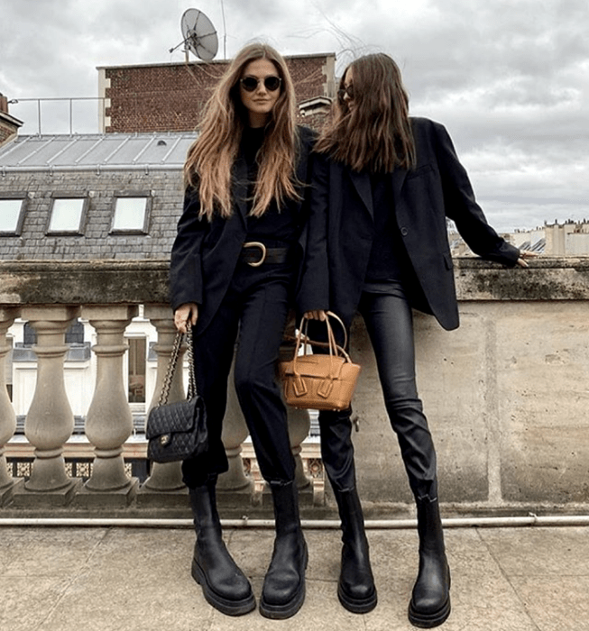 dos chicas de cabello largo usando lentes de sol, top negro de cuello alto, blazer abrigo negro, pantalones negros de vestir, pantalones de cuero, botas de plataforma negras de cuero, bolso negro de mano, bolso café de mano