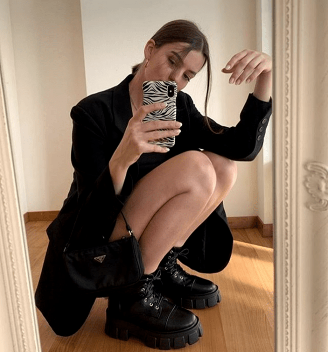 chica de cabello rubio usando un blazer oversized negro, shorts de ciclismo negros, botas de plataforma negras con suela gruesa y bolso de mano negro guess