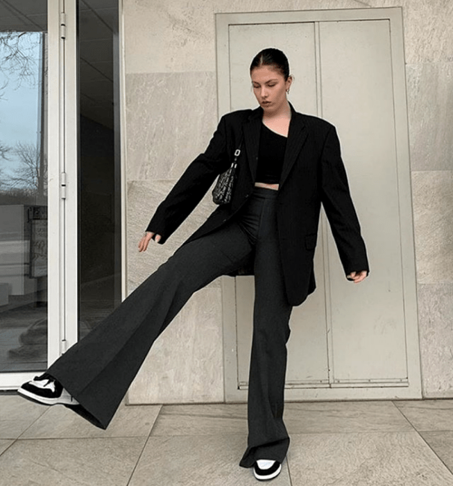 chica de cabello oscuro usando un top negro de tirantes, blazer oversized negro, pantalones de vestir acampanados oscuros, tenis blancos con negro y bolso de mano negro con blanco