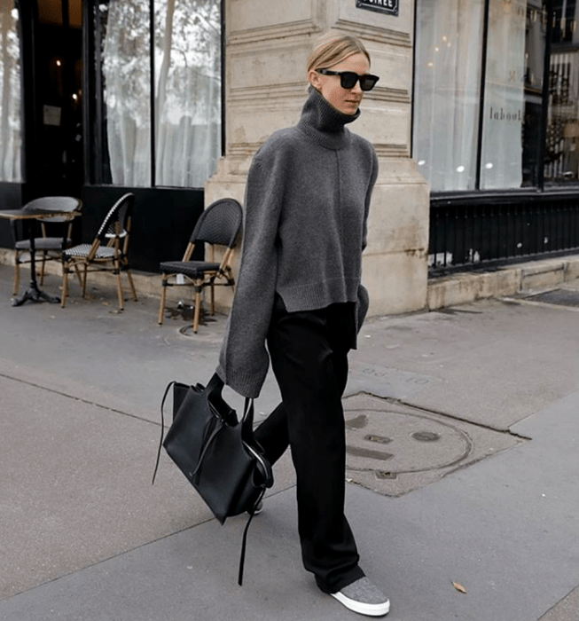 chica rubia con lentes de sol, suéter gris de cuello alto y mangas holgadas, pantalón de vestir negro, tenis grises y bolso de mano negro grande