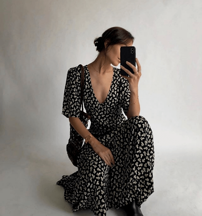 chica de cabello castaño usando un vestido negro largo con flores blancas, de cuello en V y manga 3/4 con bolso de mano negro y botines negros