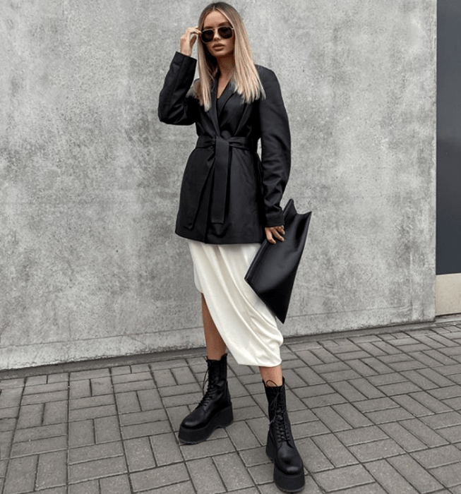 chica rubia con lentes de sol, saco blazer de cuero con cinturón, falda de satén blanca, botines negros de suela gruesa y bolso de mano clutch negro