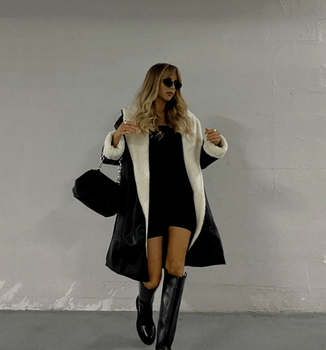 chica rubia con lentes de sol, vestido mini negro, abrigo negro con forro blanco afelpado, botas largas negras y bolso negro de mano