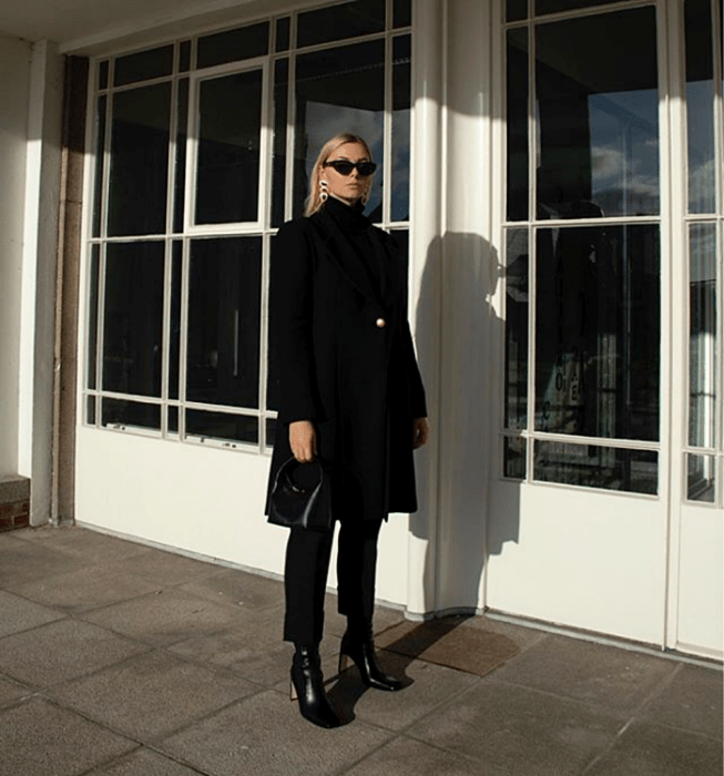 chica rubia con lentes de sol, abrigo largo  negro, suéter negro de cuello alto, pantalón de vestir negro, botines negros de tacón y bolso de mano negro