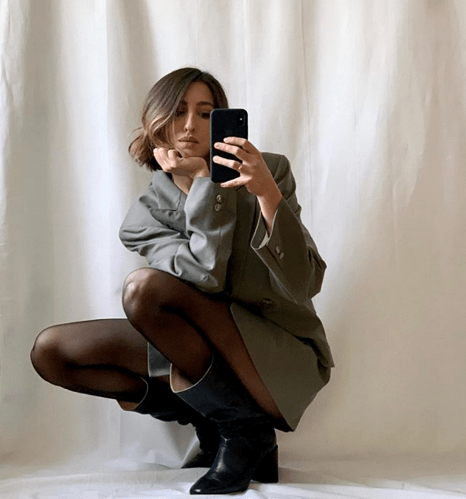 chica de cabello corto usando un blazer oversized gris, medias negras y botas largas de tacón negras