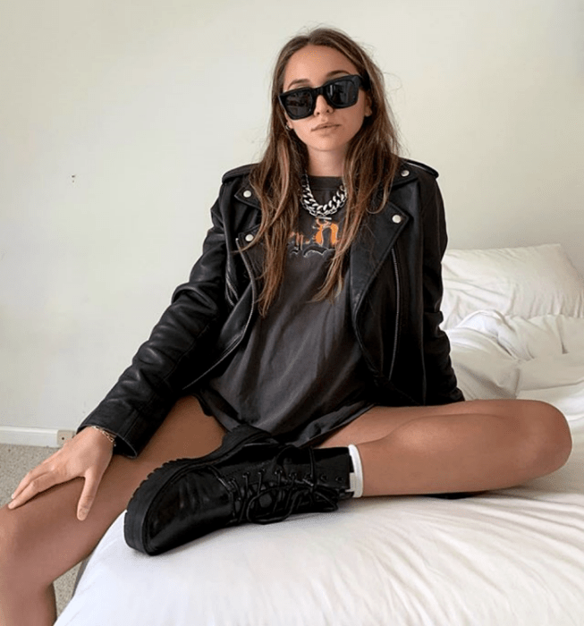 chica de cabello claro usando lentes de sol, camiseta negra con estampado gráfico, chaqueta de cuero negra, shorts negros y botines negros de suela gruesa