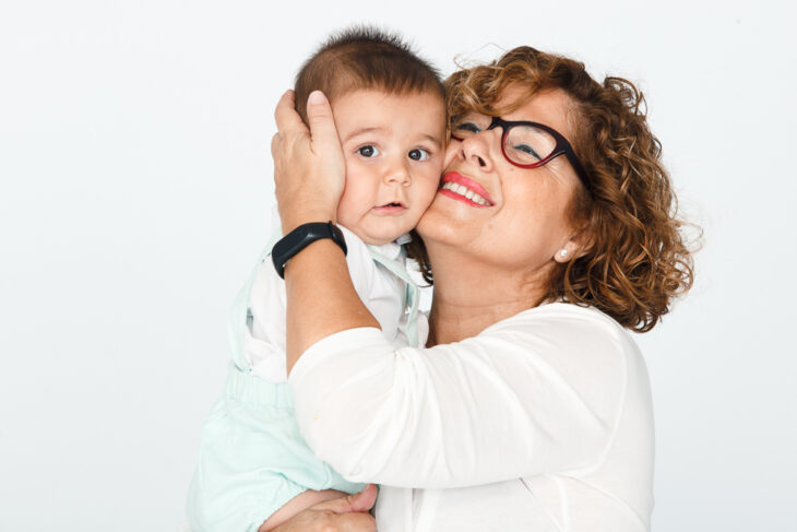 Abuela con blusa blanca y con lentes abraza a su nieto bebé en brazos 