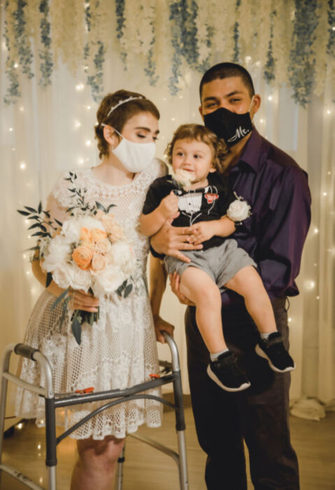 Pareja se casa en hospital gracias a enfermeras; esposos recién casados con bebé en brazos; mujer de cabello corto y castaño pelirrojo, con vestido blanco de encaje y ramo de flores, bebé rubio chino, hombre con cabello rapado, pantalón de vestir y camisa morada