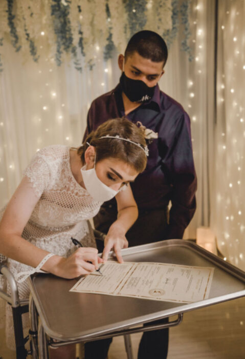 Pareja se casa en hospital gracias a enfermeras; esposos recién casados firmando acta de matrimonio, mujer de cabello corto, castaño pelirrojo, con vestido blanco de encaje, hombre con cabello rapado y camisa morada de vestid