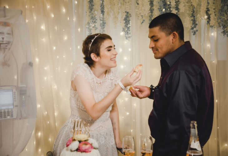 Pareja se casa en hospital gracias a enfermeras; esposos recién casados comiendo pastel de bodas, mujer de cabello corto y pelirrojo, con vestido blanco de encaje, sonriendo, hombre de cabello rapado con camisa morada de satín de vestir