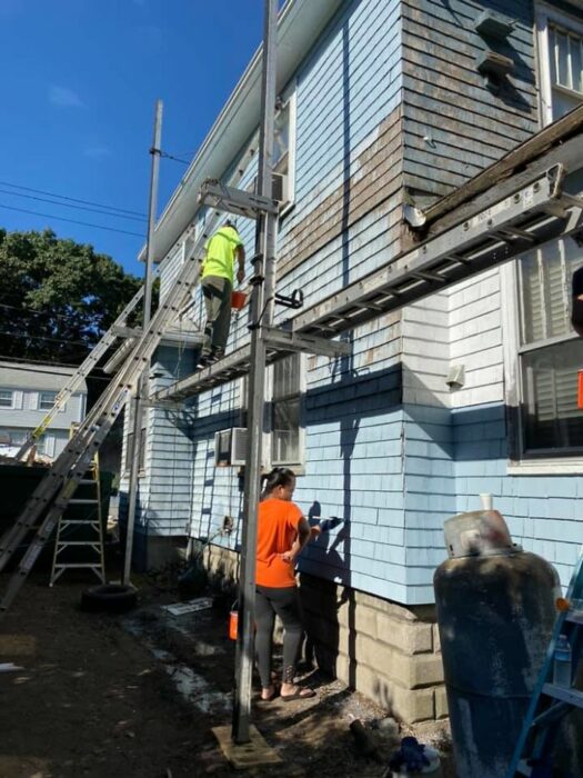 Dos personas con playeras verde neón y naranja pintando y reparando una casa de dos pisos color azul cielo 