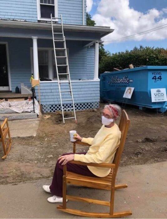 Anciana vistiendo suéter amarillo y pantalón guinda sentada en una mecedora de madera sosteniendo un vaso blanco con la mano derecha con una casa de dos pisos en el fondo color azul 