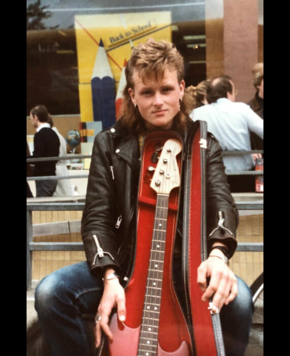 Foto vintage, fotografía antigua a color, de hombre rockero de cabello corto de arriba y largo de abajo, rubio, con bajo, chamarra de cuero negra