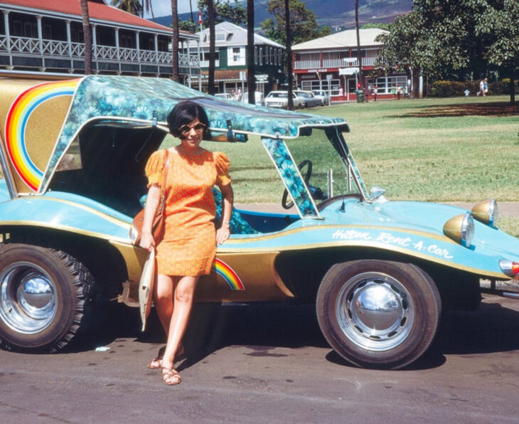 Foto vintage, fotografía antigua a color de mujer con cabello negro, corto, vestido anaranjado, lentes de sol, sandalias, recargada en un auto antiguo azul