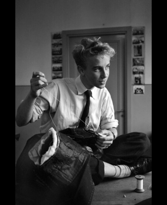 Foto vintage, fotografía antigua de hombre guapo sentado en una mesa cosiendo ropa, vestido con camisa blanca, corbata, pantalones de vestir y zapatos