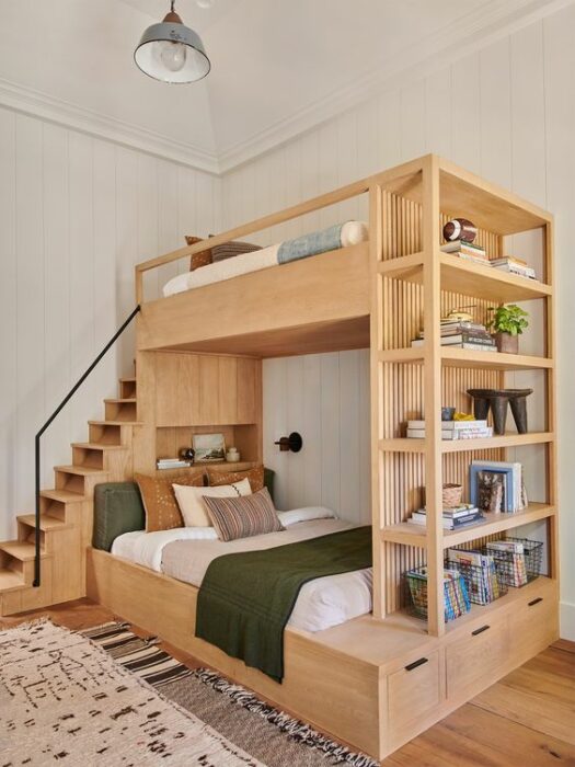 Mueble para cama de dos pisos color madera con cama matrimonial en la parte de abajo con cobijas claras y cobijes naranjas y unos escalones para subir a la planta alta