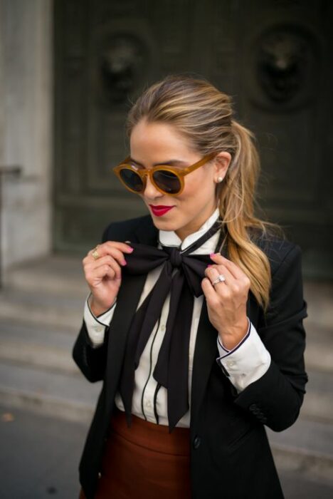 Chica rubia con coleta y lentes de sol vistiendo una falda chedrón, camisa blanca, saco negro y un moño negro en el cuello