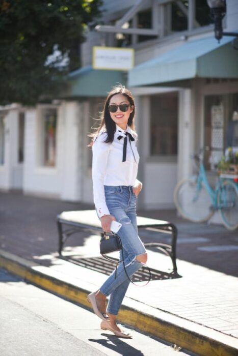 Chica delgada de cabello medio negro y lacio vistiendo jeans rotos de las rodillas, camisa blanca y moño negro en el cuello