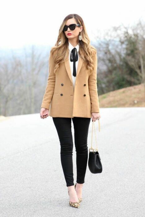 Chica rubia de cabello suelto y gafas de sol oscuras vistiendo pantalón negro, camisa blanca, abrigo beige y un moño negro en el cuello