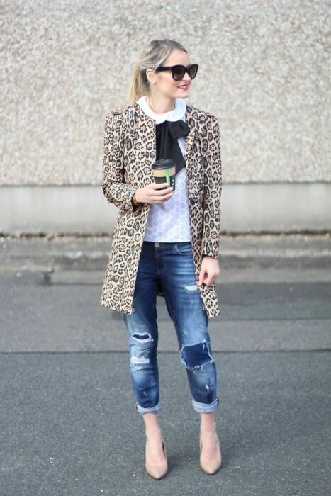 Chica rubia con coleta alta y lentes oscuros de sol vistiendo jeans oscuros, camisa blanca, saco de leopardo y moño negro en el cuello