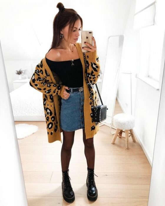 Chica usando blusa negro, mini falda denim, con medias y botines negros y un cardigan largo de estampado animal print