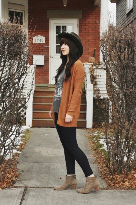 Chica usando sombrero, blusa gris, cardigan color naranja quemado, jeans negros y botines color camel