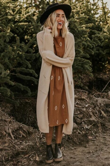 Chica usando sombrero, vestido color mostaza con cardigan largo color beige y botines color negro