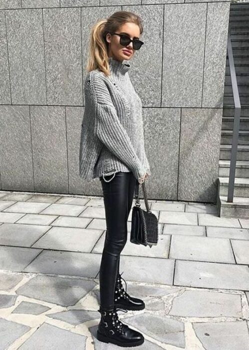 Chica usando lentes de sol, suéter oversized, leggings de vinipiel, botines negros y bolsa de mano negra