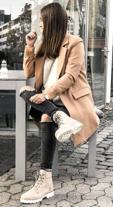 Chica usando abrigo color camel, suéter color beige y jeans negros rasgados con botines color camel 