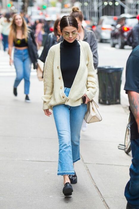 Selena Gómez caminando en la calle vistiendo pantalón de mezclilla acampanado,  zapatos negros, blusa negra de cuello y suéter largo tejido con botones beige