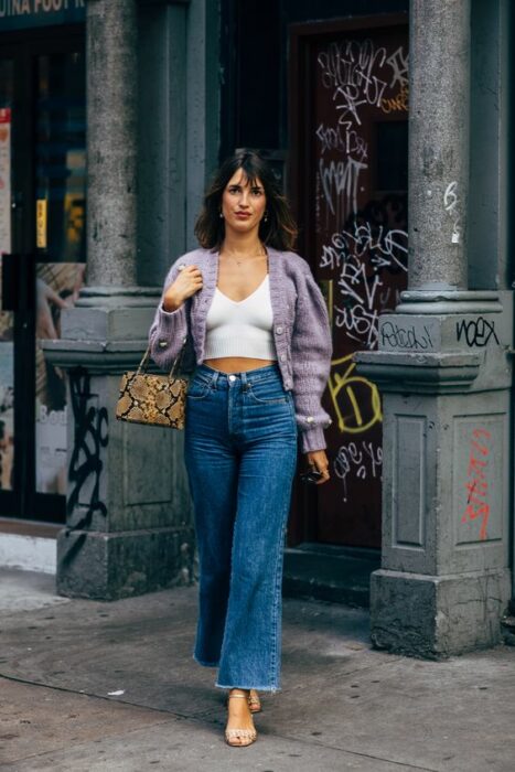 Chica parisina con jeans de campana, sandalias de tacón, blusa blanca y suéter tejido color morado con mangas anchas 