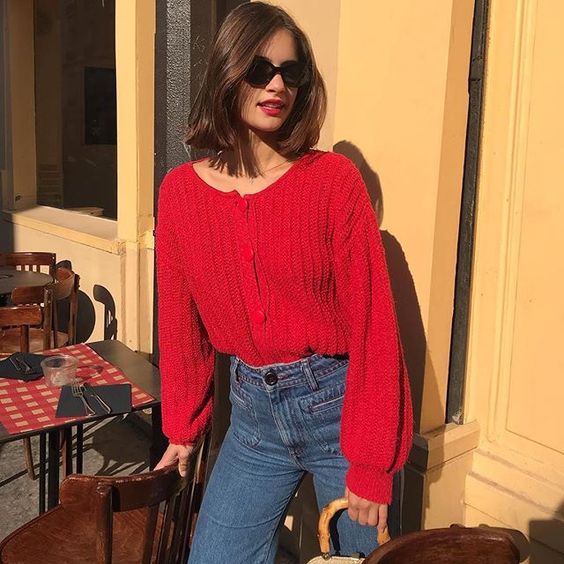 Chica delgada con melena corta, lentes de sol, labial rojo vistiendo jeans oscuros y suéter de punto rojo
