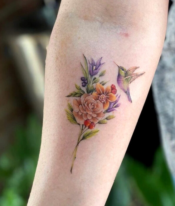 Tatuaje bonito y femenino de ave en el brazo, pájaro colibrí volando sobre flores